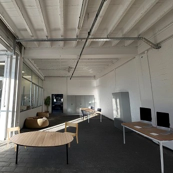 Image retouchée montrant les aménagements du mobilier dans la longueur principale du locale (à l'arrière plan,on peut voir les portes d'entrée du local). Sur la gauche on peut y voir les poufs, une table ronde. Sur la droite le long du mur, on peut voir des tables avec des oridinateurs et des armoires métalliques.