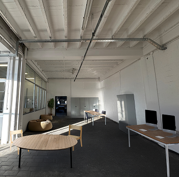 Image retouchée, montrant une vue du local, du fond du local vers la porte. Sur la gauche, il y a une table ronde et 4 chaises. Sur la droite, des tables avec des pc sont visibles.