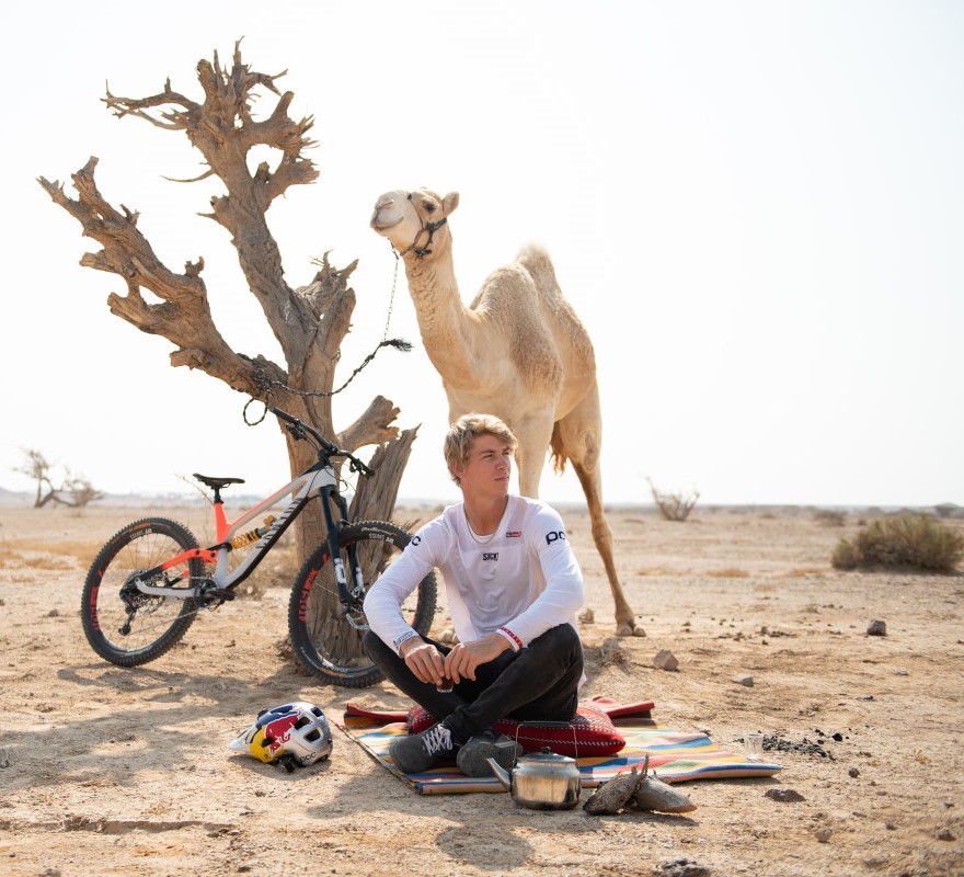 Fabio assis au pied d'un arbre accompagné d'un chameau et de son vélo, dans le désert israélien.