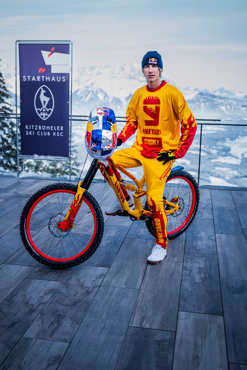 Fabio pose sur son vélo  avant de dévaler une piste de ski sur son vélo. Le décor est constiitué d'une chaine de montagne enneigée